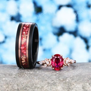 May include: A black band ring featuring a central inlay of red crushed stone and gold flakes, paired with a rose gold ring set with a central oval-cut red gemstone flanked by small clear stones on each side, both displayed on a stone surface.