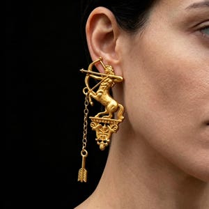 May include: Gold-coloured earrings depicting a centaur with a bow and arrow. The earrings have a detailed, three-dimensional design, with a chain and arrow pendant. The earrings are worn on a person's ear against a black backdrop.