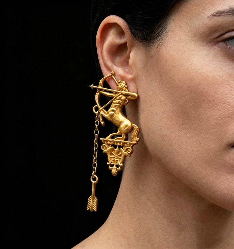 May include: Gold-coloured earrings depicting a centaur with a bow and arrow. The earrings have a detailed, three-dimensional design, with a chain and arrow pendant. The earrings are worn on a person's ear against a black backdrop.