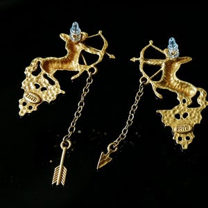May include: A pair of gold-coloured earrings featuring a centaur with a bow and arrow design. The earrings have a textured finish and dangle with a chain and arrow charm. The earrings have a post with a light blue gem.