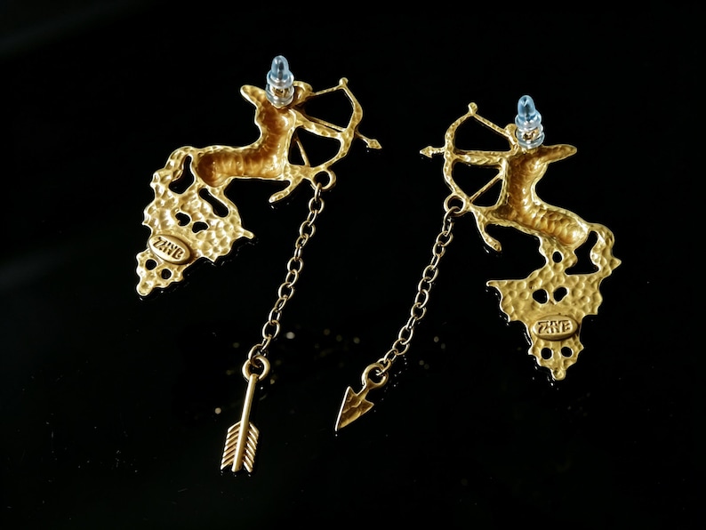 May include: A pair of gold-coloured earrings featuring a centaur with a bow and arrow design. The earrings have a textured finish and dangle with a chain and arrow charm. The earrings have a post with a light blue gem.