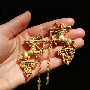 May include: Gold-coloured earrings depicting a centaur with a bow and arrow. The earrings have a detailed design with dangling chain accents and arrow charms. The earrings are vintage style and set against a dark background.