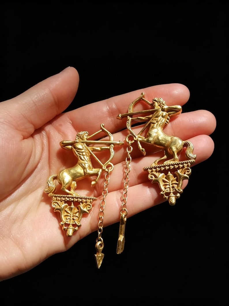 May include: Gold-coloured earrings depicting a centaur with a bow and arrow. The earrings have a detailed design with dangling chain accents and arrow charms. The earrings are vintage style and set against a dark background.