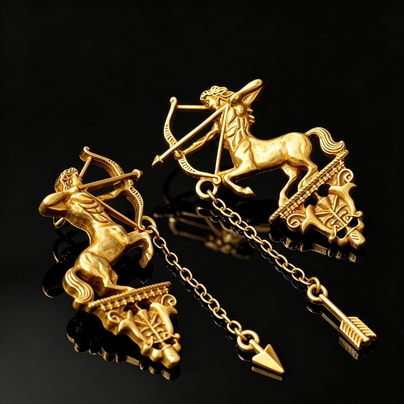 May include: Two gold-coloured Sagittarius pendant brooches. Each brooch features a centaur archer with a bow and arrow, connected by a chain to a decorative arrow pendant. The brooches are set against a black background.