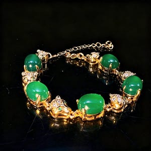 Op de afbeelding: Een goudkleurige armband met afwisselend ovale groene cabochonstenen en schakels in de vorm van panterkoppen. De panterkoppen zijn versierd met kleine heldere kristallen en groene stenen ogen. De armband heeft een goudkleurige ketting met karabijnsluiting.