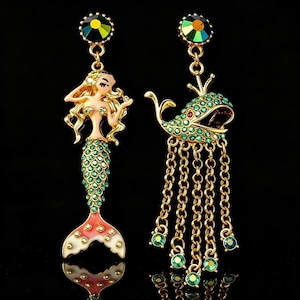May include: A pair of gold-tone dangle earrings featuring a mermaid figure with a green crystal-encrusted tail and a whale figure adorned with green crystals and multiple dangling gold chains, both topped with a round iridescent crystal stud.
