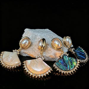 May include: Gold-toned earrings with a pearl-like oval stud and a fan-shaped drop. The drop features a mother-of-pearl or abalone shell design, outlined with small, clear crystals. The earrings are displayed on a white stone against a black background.