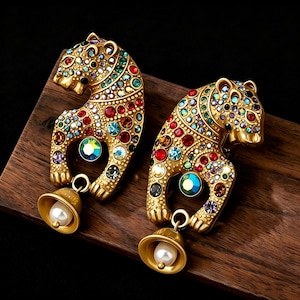 May include: A pair of gold-toned leopard-shaped earrings adorned with colourful gemstones. Each earring has a bell with a pearl. The earrings are presented on a wooden surface.