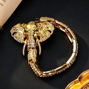 Può includere: Un bracciale color oro caratterizzato da un elemento centrale a forma di testa di elefante con grandi orecchie e zanne. La testa dell'elefante e il cinturino a maglie sono impreziositi da strass trasparenti scintillanti. Il metallo ha una finitura lucida.