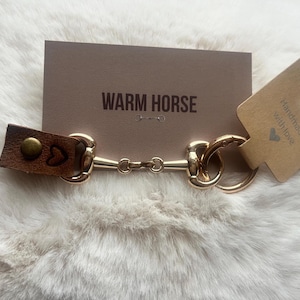 May include: A gold-colored metal horse bit keychain with a brown leather strap. The leather strap has a heart design and a brass button. A brown card with the words "WARM HORSE" is behind the keychain. A small tag says "Handmade with love".
