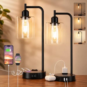 May include: Two black metal table lamps with clear cylindrical glass shades and Edison-style bulbs. Each lamp base features integrated USB ports and a power outlet, with charging cables connected to a smartphone, smartwatch, and wireless earbuds.