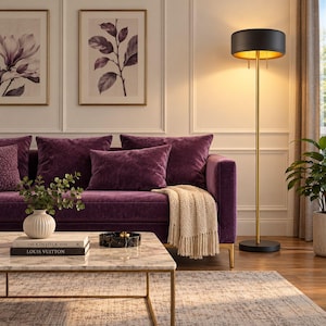 May include: A deep purple velvet sofa with a cream-colored knit throw blanket, a marble-topped coffee table with books and a vase, and a tall floor lamp with a black drum shade and gold interior, standing next to a green potted plant.