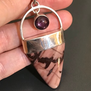 Könnte beinhalten: Ein silberner Anhänger mit einem rosa und braunen Stein in Form eines Dreiecks. Ein kleiner runder lila Stein mit einer sternförmigen Einlagerung hängt an der Oberseite des Anhängers.
