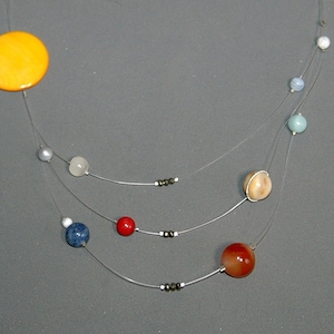 May include: A necklace featuring a large orange circular pendant and various colorful beads, strung on delicate clear wire. The beads include red, blue, and tan spheres, creating a celestial or planetary theme. The necklace is displayed against a gray background.