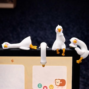 以下が含まれることがあります： 黄色い足とくちばしを持つ、白いガチョウの形をした4つのデスクアクセサリー。1羽のガチョウがぶら下がり、もう1羽が横たわり、2羽がスクリーンの端に止まっています。アクセサリーは、メモ帳のある机の上にあります。