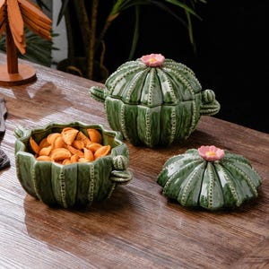 Può includere: Tre piatti da portata in ceramica verde a forma di cactus con accenti floreali rosa. Un piatto è aperto e pieno di snack. Gli altri due sono coperti. I piatti sono su una superficie di legno.