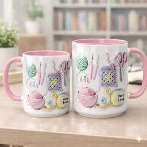 May include: Two white ceramic mugs with pink handles and interiors. The mugs are decorated with yarn balls, crochet hooks, scissors, and a crochet square. One mug has the name "Lucia" on it, and the other says "Yarn Love".