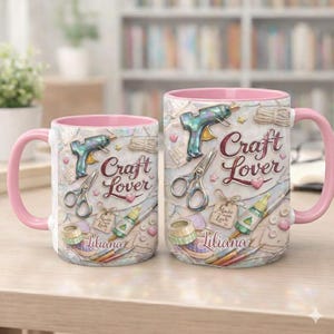 Puede incluir: Dos tazas de cerámica con asas y bordes rosas. Las tazas presentan un diseño temático de manualidades con tijeras, pistolas de pegamento y el texto "Craft Lover" y "Liliana". El fondo es de color beige claro.