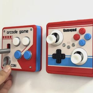 Puede incluir: Dos controladores cuadrados montados en la pared en rojo, blanco y azul. La unidad izquierda cuenta con un joystick rojo, dos botones blancos, dos botones azules y el texto arcade game y coin. La unidad derecha presenta la etiqueta gamepad, dos joysticks, una cruceta y cuatro botones de colores.