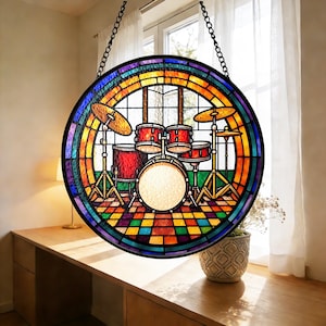 Op de afbeelding: Een rond gebrandschilderd raam met een drumstel. Het drumstel is rood en goud, met bekkens en een basdrum. Het raam heeft een regenboogrand en hangt aan een ketting.