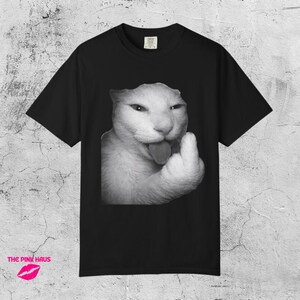 May include: Black t-shirt featuring a black and white image of a cat giving the middle finger. The cat has its tongue out. The t-shirt has a crew neck and short sleeves. The Pink Haus logo is at the bottom.
