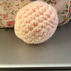 Może przedstawiać: Mały, okrągły, ręcznie robiony przedmiot na szydełku w miękkim, jasnor&oacute;żowym kolorze. Widoczna jest faktura przędzy, tworząca teksturowaną powierzchnię. Tło przedstawia wz&oacute;r kwiatowy.