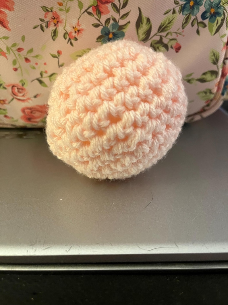 Może przedstawiać: Mały, okrągły, ręcznie robiony przedmiot na szydełku w miękkim, jasnor&oacute;żowym kolorze. Widoczna jest faktura przędzy, tworząca teksturowaną powierzchnię. Tło przedstawia wz&oacute;r kwiatowy.