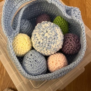 May include: A light blue crocheted basket filled with colorful crocheted eggs. The eggs are in shades of blue, yellow, pink, purple, and green. The basket has a handle and is sitting in a clear plastic container.