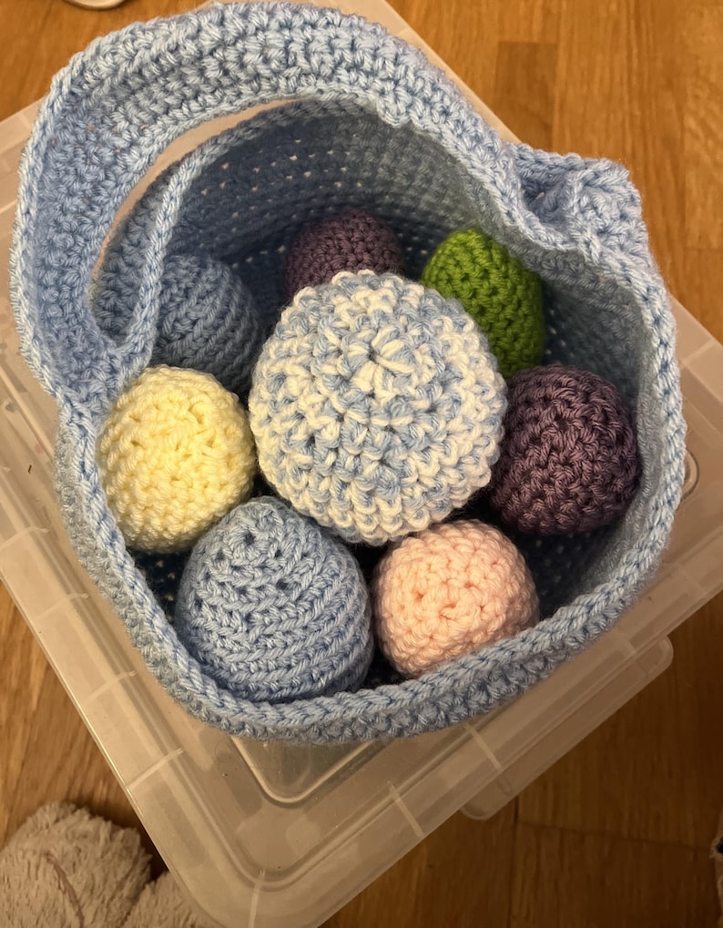 Może przedstawiać: Jasnoniebieski kosz z szydełka wypełniony kolorowymi jajkami z szydełka. Jajka są w odcieniach niebieskiego, ż&oacute;łtego, r&oacute;żowego, fioletowego i zielonego. Kosz ma uchwyt i znajduje się w przezroczystym plastikowym pojemniku.