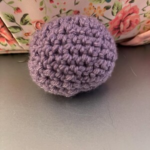 Może przedstawiać: Mała, okrągła, szydełkowa kulka w stonowanym lawendowym kolorze. Ręcznie wykonana kula ma teksturowaną powierzchnię utworzoną przez widoczne ściegi. Tło zawiera kwiatowy wz&oacute;r w kolorze r&oacute;żowym i zielonym.