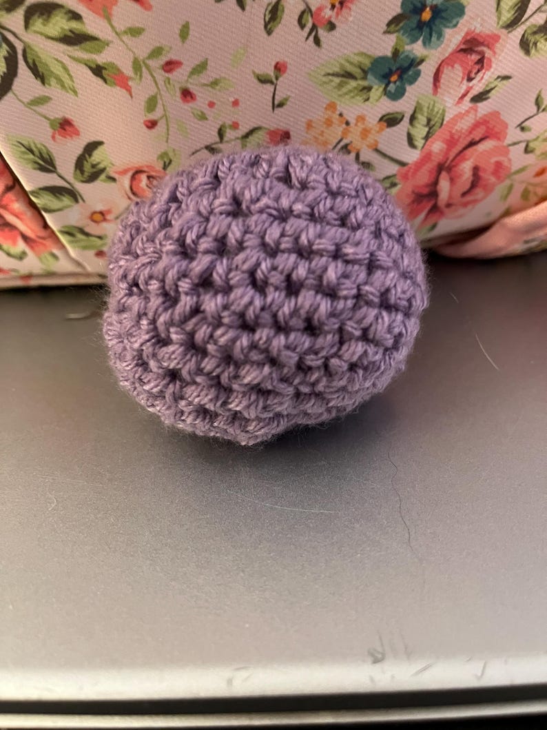 Może przedstawiać: Mała, okrągła, szydełkowa kulka w stonowanym lawendowym kolorze. Ręcznie wykonana kula ma teksturowaną powierzchnię utworzoną przez widoczne ściegi. Tło zawiera kwiatowy wz&oacute;r w kolorze r&oacute;żowym i zielonym.