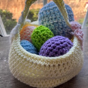 Może przedstawiać: Szydełkowy koszyk wielkanocny w miękkim ż&oacute;łtym kolorze, wypełniony kolorowymi szydełkowymi jajkami w odcieniach niebieskiego, zielonego, pomarańczowego i fioletowego. Koszyk ma pasujący ż&oacute;łty uchwyt i r&oacute;żowy detal kokardy.