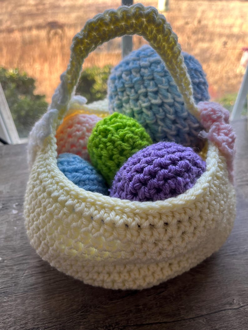 Może przedstawiać: Szydełkowy koszyk wielkanocny w miękkim ż&oacute;łtym kolorze, wypełniony kolorowymi szydełkowymi jajkami w odcieniach niebieskiego, zielonego, pomarańczowego i fioletowego. Koszyk ma pasujący ż&oacute;łty uchwyt i r&oacute;żowy detal kokardy.