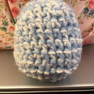 Może przedstawiać: Mały, okrągły, ręcznie robiony na szydełku przedmiot w odcieniach jasnoniebieskiego i białego. Przędza jest ciasno tkana, tworząc teksturowaną powierzchnię. Wydaje się, że jest to element dekoracyjny lub część większego projektu rzemieślniczego.