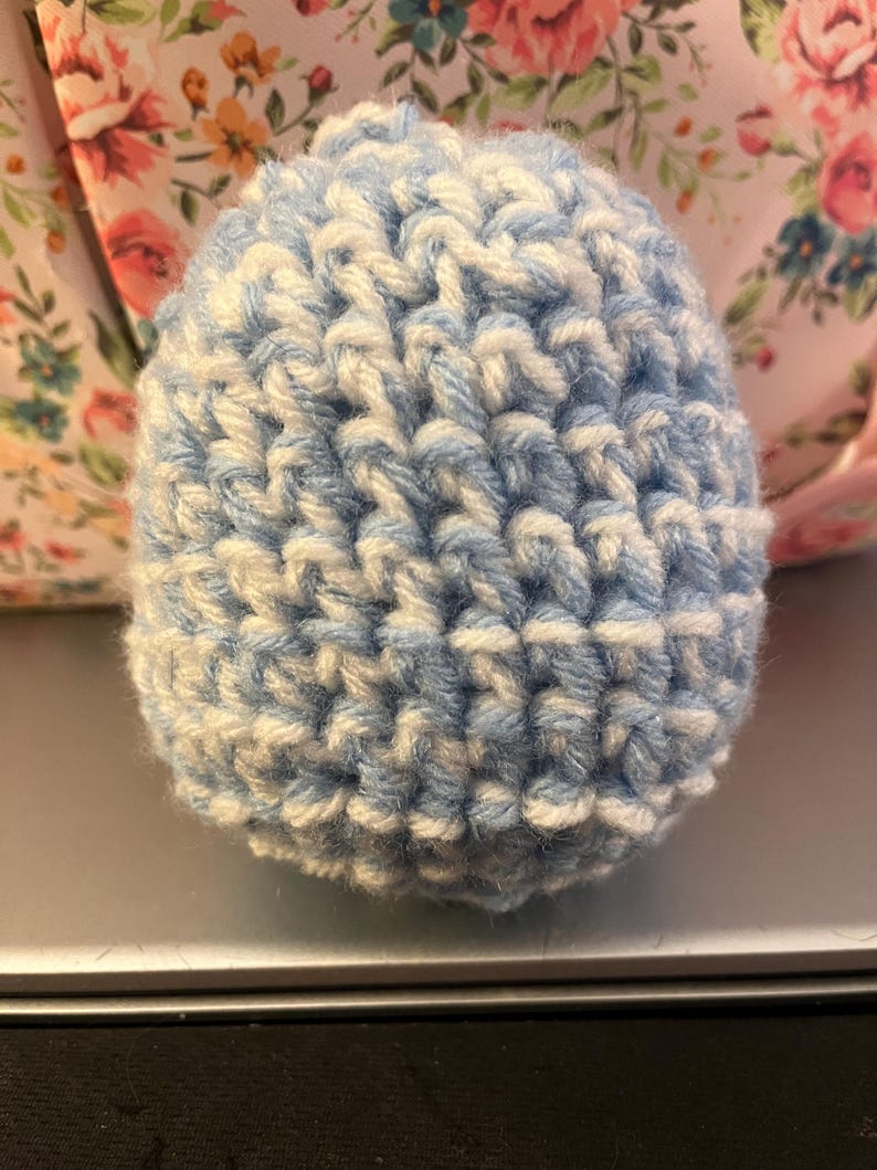 Może przedstawiać: Mały, okrągły, ręcznie robiony na szydełku przedmiot w odcieniach jasnoniebieskiego i białego. Przędza jest ciasno tkana, tworząc teksturowaną powierzchnię. Wydaje się, że jest to element dekoracyjny lub część większego projektu rzemieślniczego.