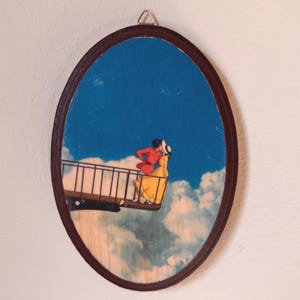 Op de afbeelding: Ovale houten wandkunst met een stel op een brug tegen een blauwe lucht met wolken. Het kunstwerk heeft een donkerbruine lijst en een metalen hanger. Het stel draagt rode en gele kleding.