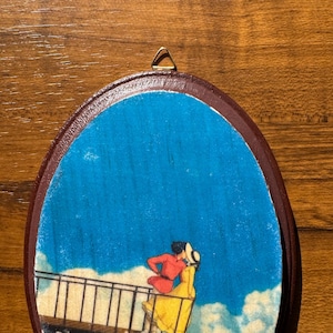 Op de afbeelding: Ovale houten wandkunst met een romantische sc&egrave;ne van twee figuren die elkaar omarmen op een balkon, tegen een blauwe lucht met wolken. De figuren zijn gekleed in rode en gele kleding. De kunst heeft een bruine lijst en een gouden hanger.