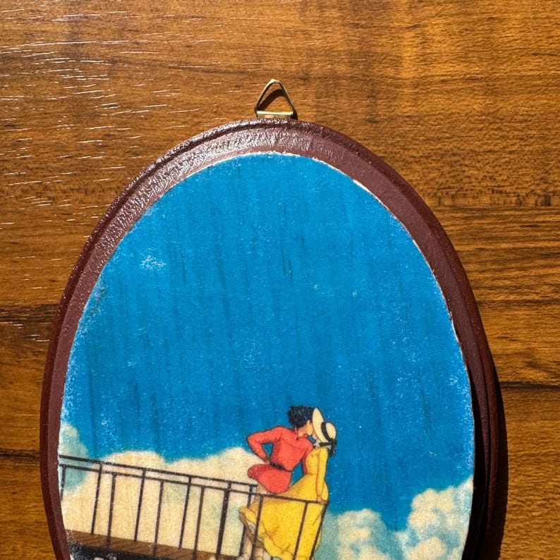 Op de afbeelding: Ovale houten wandkunst met een romantische sc&egrave;ne van twee figuren die elkaar omarmen op een balkon, tegen een blauwe lucht met wolken. De figuren zijn gekleed in rode en gele kleding. De kunst heeft een bruine lijst en een gouden hanger.