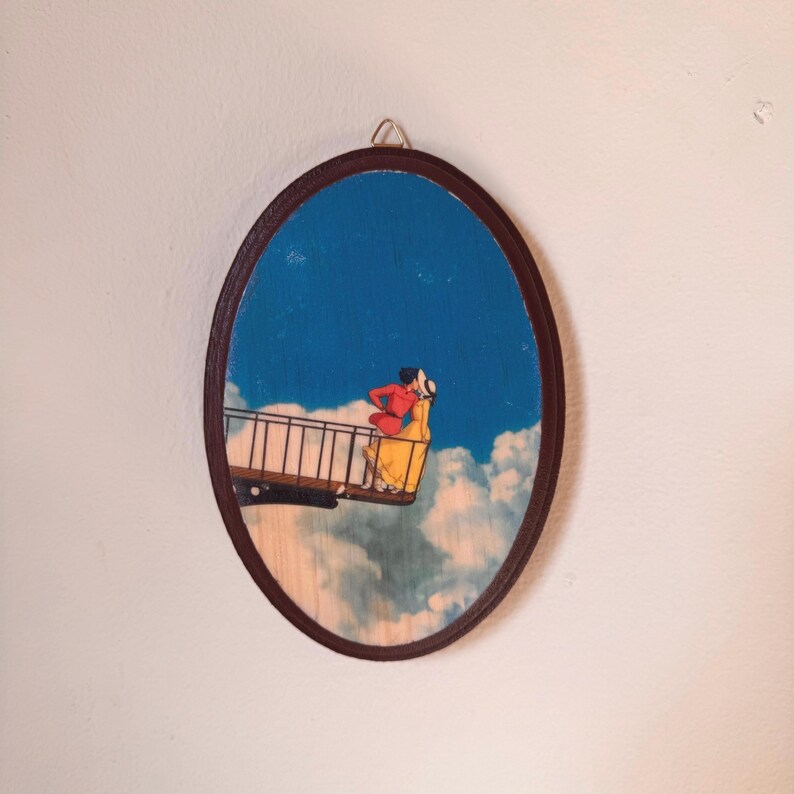 Op de afbeelding: Ovale houten wandkunst met een stel op een brug tegen een blauwe lucht met wolken. Het kunstwerk heeft een donkerbruine lijst en een metalen hanger. Het stel draagt rode en gele kleding.