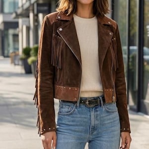 Puede incluir: Chaqueta de ante marrón con flecos en las mangas y un cuello marrón en contraste. La chaqueta tiene cierre de cremallera y detalles de tachuelas. La modelo lleva un jersey color crema y vaqueros azules.