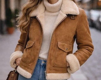 May include: A brown suede jacket with a cream-colored shearling collar and cuffs. The jacket has two front pockets and a zipper closure. The person is wearing a cream-colored turtleneck sweater and blue jeans. A brown purse is hanging from the shoulder.