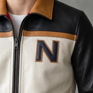 May include: A leather jacket with a color block design. The jacket features a tan collar, black and tan accents, and a large navy blue letter "N" patch. The jacket has a zipper closure.