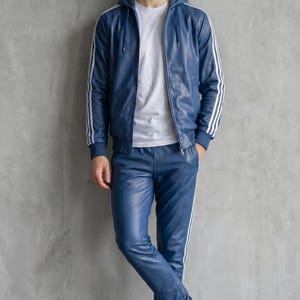 Op de afbeelding: Een blauw leren trainingspak met witte strepen op de mouwen en broekspijpen. De jas heeft een capuchon en de persoon draagt een wit t-shirt en witte sneakers. De outfit is een moderne interpretatie van een klassieke sportstijl.