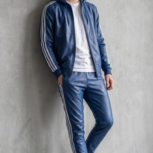 Op de afbeelding: Een blauw leren trainingspak met witte strepen op de mouwen van de jas en de broekspijpen. De jas heeft een capuchon en een rits. De persoon draagt een wit t-shirt en witte sneakers. De outfit is een sportieve, casual stijl.