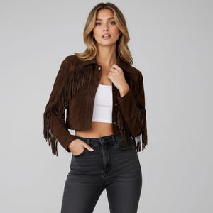 May include: A brown suede jacket with long sleeves and fringe detailing. The cropped jacket features a collar and button closure. The model is wearing a white top and black jeans.