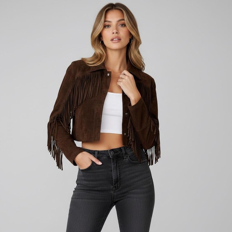May include: A brown suede jacket with long sleeves and fringe detailing. The cropped jacket features a collar and button closure. The model is wearing a white top and black jeans.