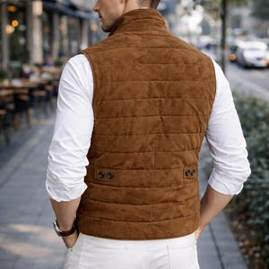 May include: A brown suede vest with a quilted design, worn over a white long-sleeve shirt and white pants. The vest has a high collar and two buttoned pockets. The person is wearing a watch and standing on a city sidewalk.