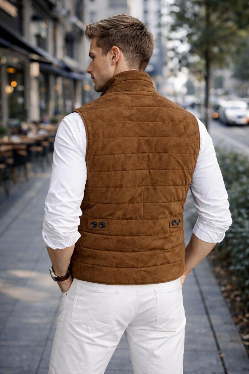 May include: A brown suede vest with a quilted design, worn over a white long-sleeve shirt and white pants. The vest has a high collar and two buttoned pockets. The person is wearing a watch and standing on a city sidewalk.
