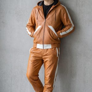 May include: A brown leather tracksuit with white stripes and accents. The jacket has a hood and a zipper closure. The pants have a drawstring waist. The outfit is worn with white sneakers. The tracksuit is made of a textured leather material.