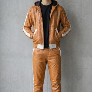 May include: A brown leather tracksuit with white stripes on the sides. The tracksuit includes a hooded jacket and matching pants. The person is wearing a black t-shirt and white sneakers. The outfit has a textured pattern.